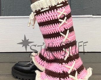 May include: Crocheted leg warmers in pink and brown stripes with cream-colored trim and lace-up details. The leg warmers are worn over black shoes with a chunky sole. The overall aesthetic is a mix of handmade and modern fashion.