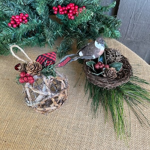 May include: Two Christmas ornaments, one is a brown twig ball with red berries and a plaid ribbon, the other is a bird in a brown nest with red berries and pine needles.