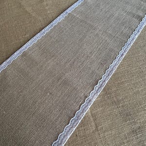 May include: A rectangular burlap table runner with white lace trim along the edges. The runner is a natural tan color and is displayed on a textured surface.