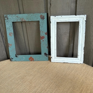 May include: Two rectangular picture frames. One is painted teal with red and brown paint splatters. The other is white with a distressed finish. Both frames are empty and ready for photos or artwork. The frames are on a burlap surface.