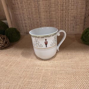 May include: White ceramic mug with a green and gold trim. The mug features a Christmas tree design with a red and white nutcracker figure.