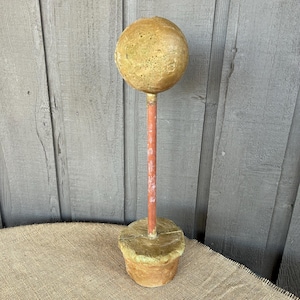 May include: A decorative, handcrafted garden ornament. It features a round, textured, golden-yellow sphere atop a reddish-orange stem, set in a weathered, cylindrical base. The piece has a rustic, aged appearance, suggesting outdoor use.