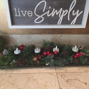May include: A rectangular centerpiece with faux greenery, red berries, pinecones, and four white candle lights. The centerpiece is set on a marble surface, with a wooden sign that reads "live Simply" in the background.