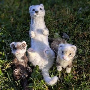 May include: Three handcrafted ferret figures on grass. One is white and grey, standing up. The other two are brown and tan, in a playful pose. The figures are made of soft, textured material, with detailed features.