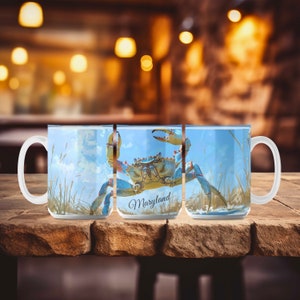 May include: White ceramic mug featuring a blue crab illustration with the word "Maryland" in script. The crab is set against a blue sky and grassy background. The mug has a large handle.