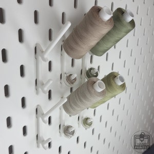 Könnte beinhalten: Weiße Lochwand mit weißen Garnrollenhaltern, die Garnrollen in Beige- und Grüntönen halten. Die Lochwand hat ein Gitter aus ovalen Löchern. In der unteren rechten Ecke befindet sich ein "Hand Printed"-Logo.