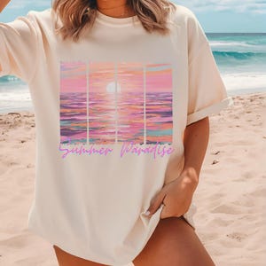 Puede incluir: Camiseta crema de gran tamaño con un gráfico de puesta de sol. El diseño muestra una colorida puesta de sol sobre el mar, con tonos rosas, naranjas y morados. Las palabras "Summer Paradise" están escritas en cursiva debajo.