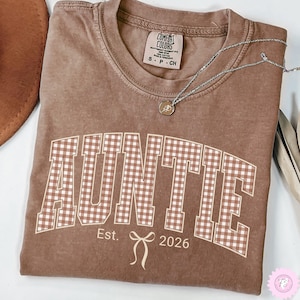 May include: A brown short-sleeve t-shirt with a crew neckline. The front features the word AUNTIE in a white-outlined brown and white gingham pattern, with the text Est. 2026 and a small bow graphic centered below in white ink.