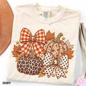 May include: An ivory short-sleeve t-shirt featuring a graphic print of two pumpkins, one with a floral pattern and one with a leopard print, accented by a large orange gingham bow, a white polka dot bow, autumn leaves, and small gold stars.