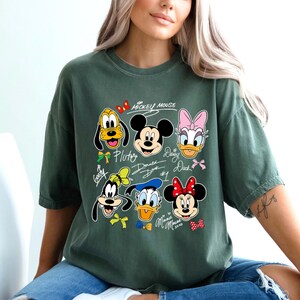 May include: Green t-shirt featuring Mickey Mouse and friends. The design includes Pluto, Mickey, Daisy Duck, Donald Duck, Goofy, and Minnie Mouse. The text "Mickey Mouse" is above the characters.