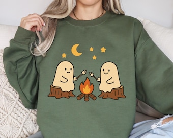 May include: Olive green sweatshirt featuring a whimsical illustration of two ghost-like figures roasting marshmallows over a campfire. The design includes a crescent moon and stars, creating a cozy, outdoorsy theme.