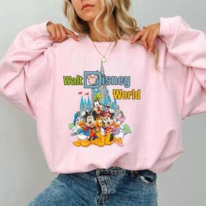 Puede incluir: Sudadera rosa claro con un gr&aacute;fico colorido de personajes de Disney, incluyendo Mickey Mouse, Minnie Mouse, Donald Duck y Goofy, frente a un castillo. Las palabras "Walt Disney World" est&aacute;n impresas encima de los personajes en verde, azul y amarillo.