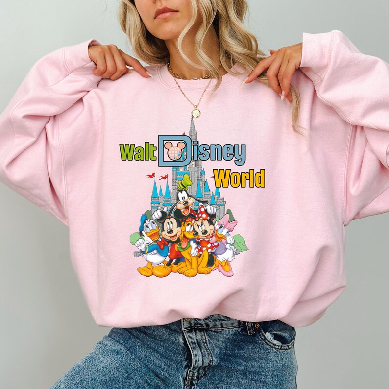 Puede incluir: Sudadera rosa claro con un gr&aacute;fico colorido de personajes de Disney, incluyendo Mickey Mouse, Minnie Mouse, Donald Duck y Goofy, frente a un castillo. Las palabras "Walt Disney World" est&aacute;n impresas encima de los personajes en verde, azul y amarillo.