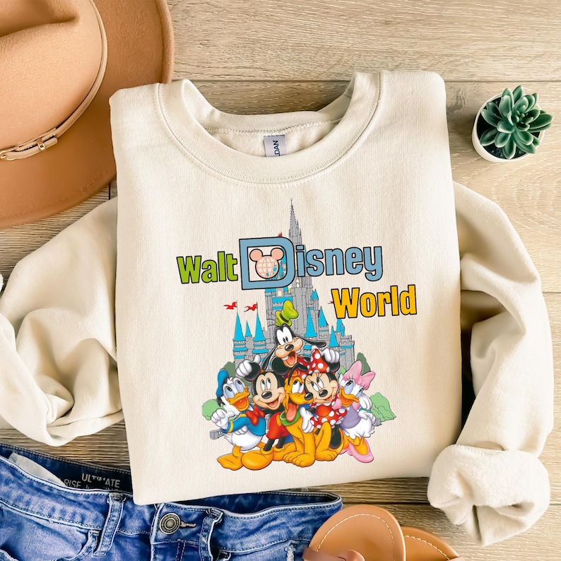 Puede incluir: Sudadera color crema con el texto "Walt Disney World" y un gr&aacute;fico de personajes de Disney y el castillo de Cenicienta. El dise&ntilde;o incluye Mickey Mouse, Pato Donald, Goofy, Minnie Mouse y Daisy Duck.