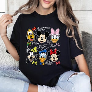 May include: Black t-shirt featuring a colorful graphic of Disney characters including Mickey Mouse, Pluto, Daisy Duck, and Minnie Mouse. The design includes character signatures and the text "Mickey Mouse".