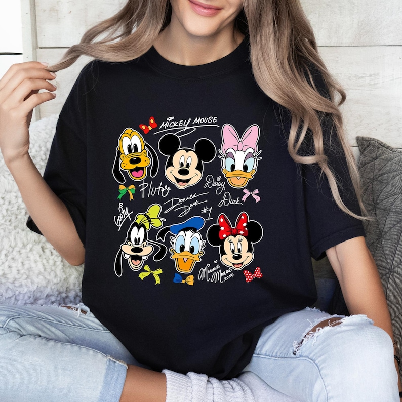 May include: Black t-shirt featuring a colorful graphic of Disney characters including Mickey Mouse, Pluto, Daisy Duck, and Minnie Mouse. The design includes character signatures and the text "Mickey Mouse".