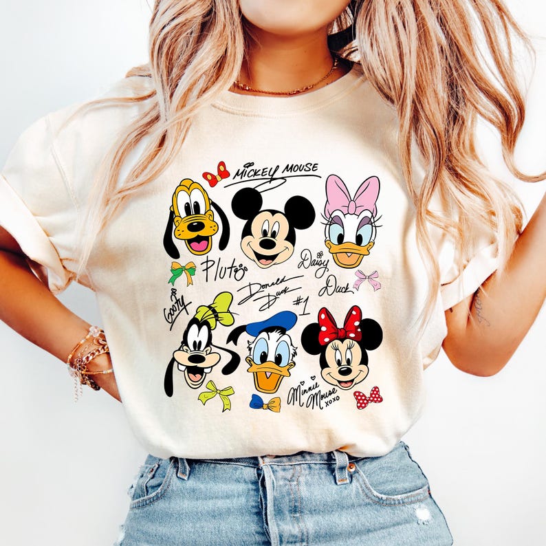 May include: Cream-colored t-shirt featuring cartoon illustrations of Mickey Mouse, Pluto, Goofy, Donald Duck, Daisy Duck, and Minnie Mouse. The characters' names are written below them in a handwritten style. The words "Mickey Mouse" are written above the characters.