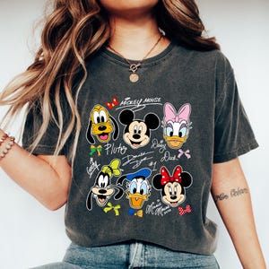 May include: Dark gray t-shirt featuring a graphic of Mickey Mouse and friends. The design includes Pluto, Mickey, Daisy Duck, Donald Duck, Goofy, and Minnie Mouse. The words "Mickey Mouse" are at the top of the design.