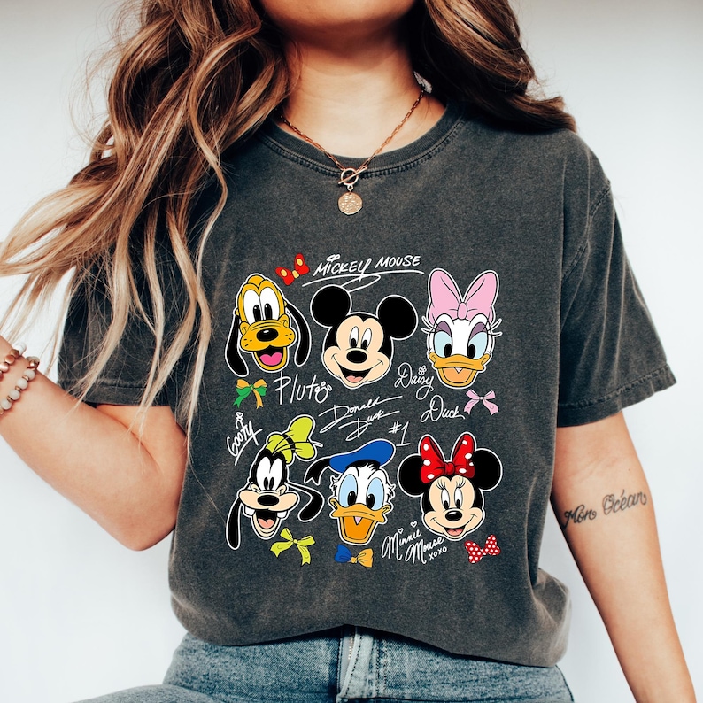 May include: Dark gray t-shirt featuring a graphic of Mickey Mouse and friends. The design includes Pluto, Mickey, Daisy Duck, Donald Duck, Goofy, and Minnie Mouse. The words "Mickey Mouse" are at the top of the design.