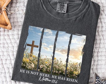 May include: A dark gray t-shirt with a distressed graphic of a wooden cross in a field of flowers. The graphic is surrounded by a white border and the text "He is not here. He has risen. Matthew 28:6" is printed below the graphic.