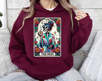 May include: A maroon hooded sweatshirt featuring a graphic print of a skeleton in a floral robe holding a coffee mug. The design is framed like a tarot card with colorful flowers and the text TM and THE MOM printed in black lettering.