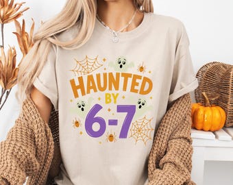 May include: Beige t-shirt with the text "HAUNTED BY 6-7" in orange and purple. The design includes cartoon ghosts, spiderwebs, and spiders, perfect for Halloween. The shirt is a casual, short-sleeved style.