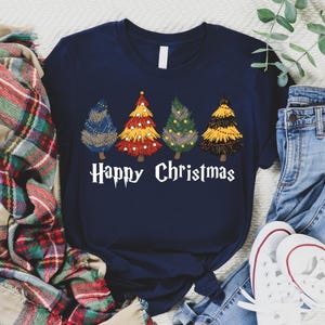 May include: Navy blue t-shirt featuring four stylized Christmas trees in blue, red, green, and yellow, each with unique decorative elements. The words "Happy Christmas" are printed in white below the trees. The shirt is paired with jeans and sneakers.