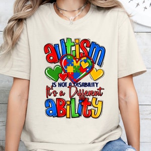 May include: Cream-colored t-shirt with a colorful graphic design. The design includes the word "Autism" in red and blue, along with the phrase "is not a disability, it's a different ability." A heart-shaped puzzle piece is in the center.