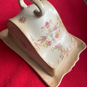 May include: A vintage ceramic cheese dish with a floral design and a handle. The dish has a lid with a small hole in the top.