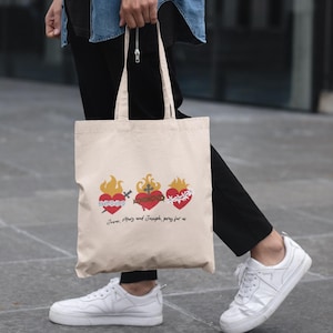 Puede incluir: Una bolsa de lona beige con tres corazones rojos con llamas y detalles florales. El texto "Jesús, María y José, reza por nosotros" está impreso en la bolsa.