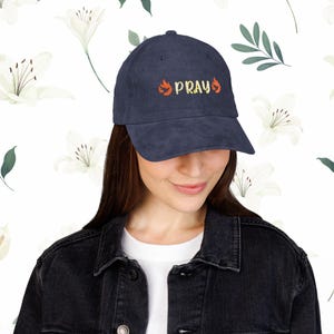 Puede incluir: Gorra de béisbol de pana azul marino con la palabra "PRAY" bordada en amarillo y naranja. La gorra presenta una visera curva y un diseño clásico de seis paneles. El texto va acompañado de gráficos de llamas.
