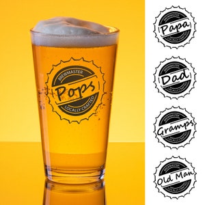 Puede incluir: Un vaso de pinta de vidrio transparente con un líquido amarillo en su interior. El vaso tiene una pegatina en blanco y negro que dice "Brewmaster Pops Locally Crafted".