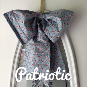 Peut inclure: Un grand nœud décoratif en tissu bleu avec un motif d'étoiles rouges et blanches. Le nœud est suspendu à un miroir encadré de blanc. Le mot "Patriotic" est écrit sur le miroir.