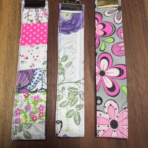 May include: Three fabric key fobs with metal key rings. The fobs feature different patterns: pink polka dots and floral, purple floral, and pink flowers on gray. Each fob is rectangular and sewn with a metal ring.