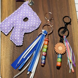 Op de afbeelding: Drie sleutelhangers met gepersonaliseerde details. Eén heeft een paarse stipjesletter, een andere heeft de naam "MORGAN" met kleurrijke kralen, en de derde toont "MAR" met een bloemcharm. Elke sleutelhanger heeft een ander ontwerp.