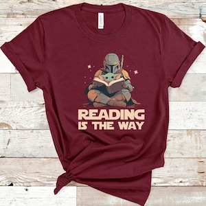 May include: Burgundy t-shirt featuring a cartoon illustration of a character in armor reading a book with a small green creature. The text "READING IS THE WAY" is below the illustration. The shirt is made of a soft material.