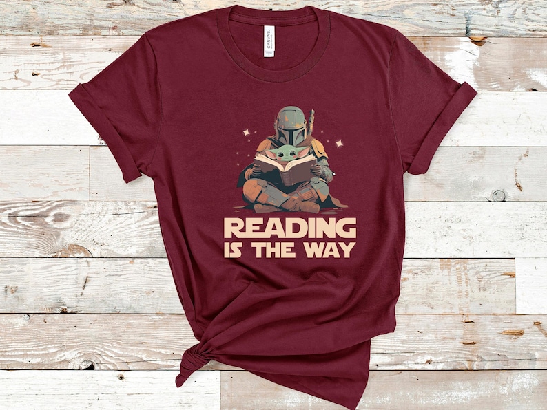 May include: Burgundy t-shirt featuring a cartoon illustration of a character in armor reading a book with a small green creature. The text "READING IS THE WAY" is below the illustration. The shirt is made of a soft material.