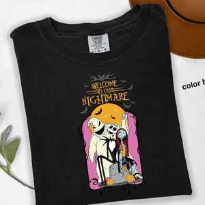 Puede incluir: Camiseta negra con un gr&aacute;fico de "Pesadilla antes de Navidad". El dise&ntilde;o incluye a Jack Skellington, Sally y Oogie Boogie, con el texto "Welcome to our Nightmare". El dise&ntilde;o est&aacute; sobre un fondo rosa con una luna amarilla.