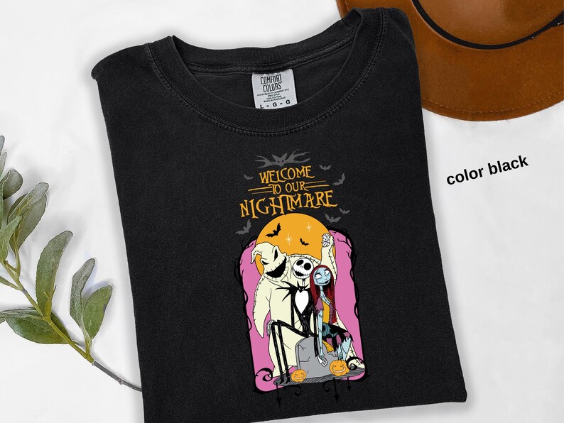 Puede incluir: Camiseta negra con un gr&aacute;fico de "Pesadilla antes de Navidad". El dise&ntilde;o incluye a Jack Skellington, Sally y Oogie Boogie, con el texto "Welcome to our Nightmare". El dise&ntilde;o est&aacute; sobre un fondo rosa con una luna amarilla.