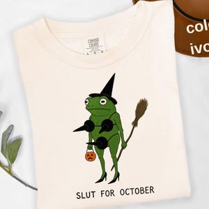 May include: Ivory-colored t-shirt featuring a cartoon frog dressed as a witch, holding a broom and a jack-o'-lantern. The text "SLUT FOR OCTOBER" is printed below the frog. The shirt is a Comfort Colors brand.