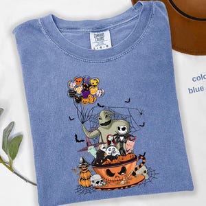 Pode incluir: Camiseta azul jeans com um gr&aacute;fico tem&aacute;tico de Halloween. O design inclui personagens de O Estranho Mundo de Jack, morcegos e uma ab&oacute;bora. A camisa tem gola redonda e mangas curtas. O texto "color blue jean" &eacute; vis&iacute;vel.
