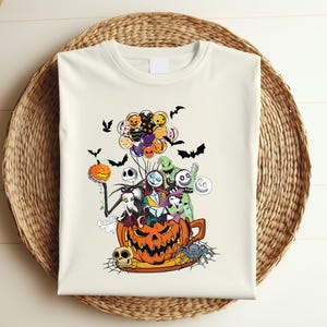 K&ouml;nnte beinhalten: Cremefarbenes T-Shirt mit einem farbenfrohen Halloween-Motiv. Das Design zeigt Charaktere aus The Nightmare Before Christmas, Flederm&auml;use, K&uuml;rbisse und Luftballons. Das T-Shirt wird auf einem Weidenkorb pr&auml;sentiert.