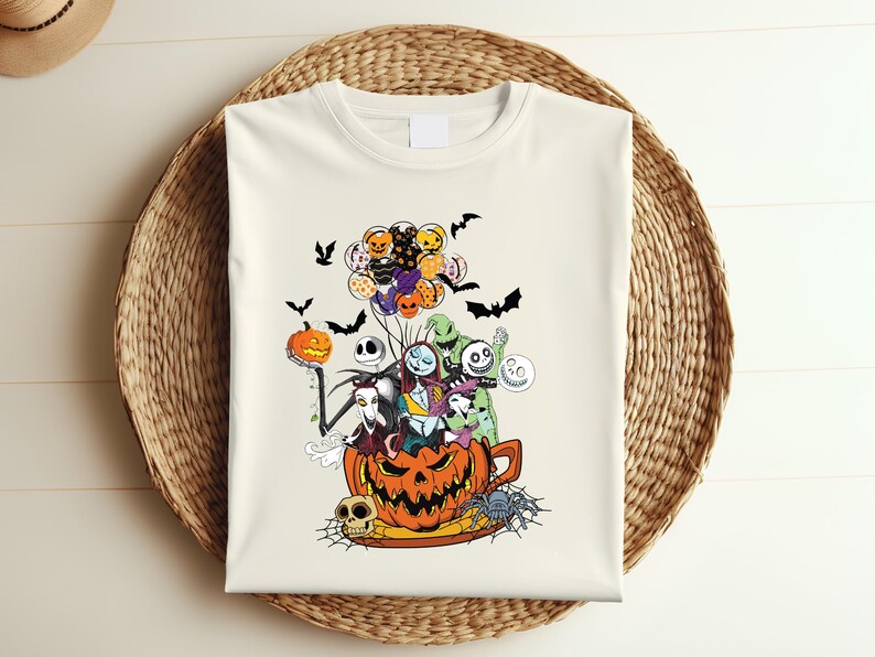 K&ouml;nnte beinhalten: Cremefarbenes T-Shirt mit einem farbenfrohen Halloween-Motiv. Das Design zeigt Charaktere aus The Nightmare Before Christmas, Flederm&auml;use, K&uuml;rbisse und Luftballons. Das T-Shirt wird auf einem Weidenkorb pr&auml;sentiert.