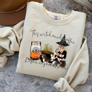 May include: Cream-colored sweatshirt with a witch-themed graphic. The design includes a witch with a black hat, a jar of orange liquid, a cauldron, and the text "This witch needs coffee before any hocus pocus."