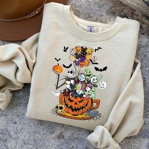 K&ouml;nnte beinhalten: Beiges Sweatshirt mit einem Halloween-Motiv. Das Design zeigt einen K&uuml;rbis, Charaktere, Flederm&auml;use und Luftballons. Die Grafik ist in Orange, Schwarz, Lila und Wei&szlig; gehalten.