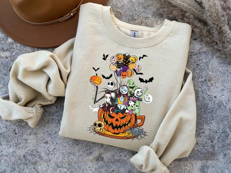 K&ouml;nnte beinhalten: Beiges Sweatshirt mit einem Halloween-Motiv. Das Design zeigt einen K&uuml;rbis, Charaktere, Flederm&auml;use und Luftballons. Die Grafik ist in Orange, Schwarz, Lila und Wei&szlig; gehalten.