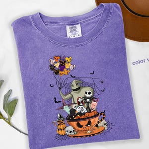 Pode incluir: Uma camiseta Comfort Colors violeta com um gr&aacute;fico tem&aacute;tico de Halloween. O design inclui personagens como Oogie Boogie e Jack Skellington, junto com morcegos, aranhas e uma x&iacute;cara de ch&aacute; em forma de ab&oacute;bora. A camisa tem gola redonda e mangas curtas.