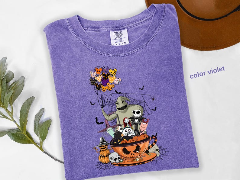 Pode incluir: Uma camiseta Comfort Colors violeta com um gr&aacute;fico tem&aacute;tico de Halloween. O design inclui personagens como Oogie Boogie e Jack Skellington, junto com morcegos, aranhas e uma x&iacute;cara de ch&aacute; em forma de ab&oacute;bora. A camisa tem gola redonda e mangas curtas.