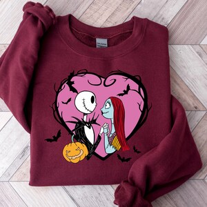 May include: A burgundy sweatshirt with a graphic of Jack Skellington and Sally from The Nightmare Before Christmas, inside a pink heart with bats. A carved pumpkin is at the bottom. The design is centered on the front.