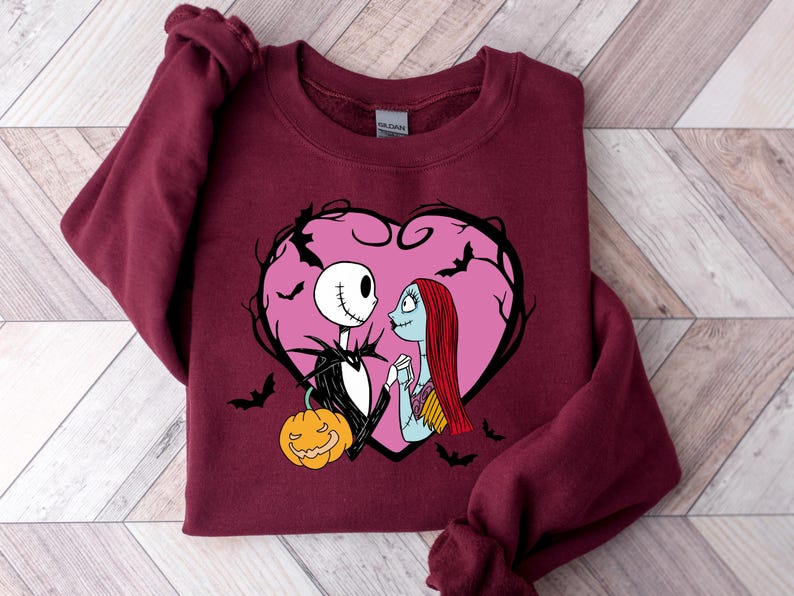 May include: A burgundy sweatshirt with a graphic of Jack Skellington and Sally from The Nightmare Before Christmas, inside a pink heart with bats. A carved pumpkin is at the bottom. The design is centered on the front.